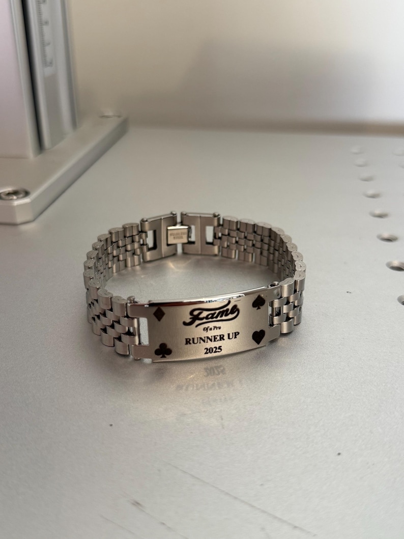 Puede incluir: Una pulsera de metal plateado con una placa rectangular grabada con la palabra "Fame", el texto "of a Pro", "Runner Up" y el a&ntilde;o "2025". La placa tambi&eacute;n presenta s&iacute;mbolos de palos de cartas. La pulsera tiene un dise&ntilde;o de eslabones.