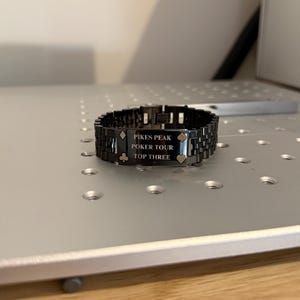 Puede incluir: Una pulsera de metal negro con el texto "PIKES PEAK POKER TOUR TOP THREE" grabado en una placa rectangular. La pulsera presenta un dise&ntilde;o de eslabones y est&aacute; sobre una superficie plateada. Tambi&eacute;n hay s&iacute;mbolos de p&oacute;quer grabados.