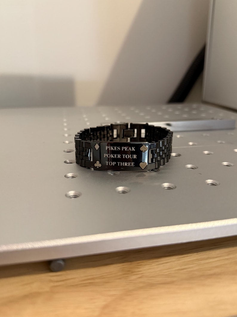 Puede incluir: Una pulsera de metal negro con el texto "PIKES PEAK POKER TOUR TOP THREE" grabado en una placa rectangular. La pulsera presenta un dise&ntilde;o de eslabones y est&aacute; sobre una superficie plateada. Tambi&eacute;n hay s&iacute;mbolos de p&oacute;quer grabados.