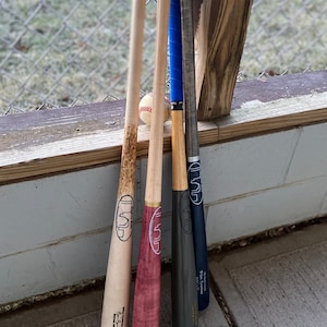 May include: Four wooden baseball bats with a baseball resting on top of them. The bats are all different colors and have a city skyline logo on them. The bats are leaning against a wooden fence.