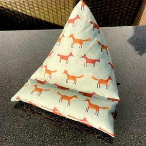 Könnte beinhalten: Ein hellgrüner, dreieckiger Buchständer aus Stoff mit einem sich wiederholenden Muster aus orangefarbenen Füchsen. Der Ständer hat eine schwarze Basis. Die Füchse sind in einem einfachen, stilisierten Design dargestellt.
