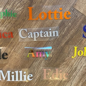 May include: A collection of iron-on vinyl names in various colours, including green, orange, red, white, black, rainbow, blue, yellow, and gold. The names include Sophie, Lottie, Jessica, Captain, Sam, Johnny, Jude, Amy, Millie, and Edie.
