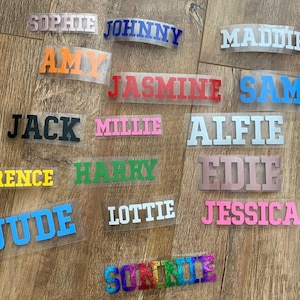 May include: A collection of iron-on name labels in various colours, including pink, blue, green, yellow, white, black, and red. The names include Sophie, Johnny, Maddie, Amy, Jasmine, Sam, Jack, Millie, Alfie, Florence, Harry, Edie, Jude, Lottie, Jessica, and Sonnie.