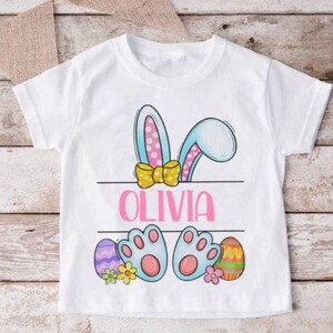 Peut inclure: T-shirt blanc avec un lapin de dessin animé qui regarde par-dessus une pancarte avec le nom "Olivia" imprimé dessus. Le lapin a des oreilles bleues à pois roses, un nœud jaune et des pieds bleus. Il y a des œufs de Pâques colorés et des fleurs sur le t-shirt.