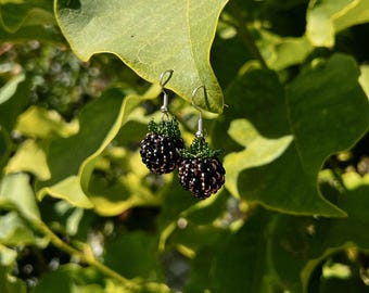 May include: A pair of dangle earrings shaped like blackberries, crafted from small dark purple and black beads with green beaded leaves at the top. Each earring features a silver-toned metal hook for attachment, mimicking the texture of fruit.