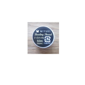 Könnte beinhalten: Ein runder Behälter in Schwarz und Weiß mit dem Text "MIYUKI Beading Thread" in goldenen Buchstaben. Der Behälter trägt außerdem den Text "ビーズステッチ糸 50m (55yds.) フィルム:PET" in Japanisch und ein Recycling-Symbol.