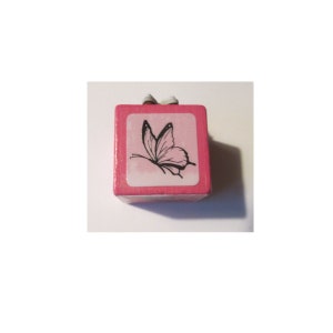 Könnte beinhalten: Ein kleiner, rosa Holzblock mit einem Schmetterlingsdesign. Der Schmetterling ist schwarz und weiß, vor einem rosa Hintergrund. Oben ist ein kleines, weißes Band befestigt. Es handelt sich wahrscheinlich um einen dekorativen Stempel.