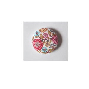 Könnte beinhalten: Ein runder Knopf mit floralem Muster. Der Knopf hat einen weißen Hintergrund mit roten, rosa, gelben und blauen Blumen. Der Knopf hat ein detailliertes Design mit Blättern und Stielen.