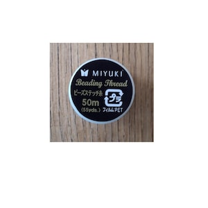 Könnte beinhalten: Eine runde Spule mit schwarzem Miyuki-Perlengarn mit weißer und goldener Schrift. Das Etikett zeigt den Markennamen, die Produktart, eine Länge von 50m (55yds.) und ein Recyclingsymbol. Die Spule hat einen silberfarbenen Rand und eine schwarze Oberseite.