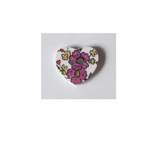 Könnte beinhalten: Ein weißer herzförmiger Knopf mit einem Blumenmuster. Das Muster zeigt rosa und gelbe Blumen auf weißem Hintergrund.