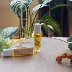 May include: Two glass roll-on bottles filled with yellow liquid, one labeled "Focus." The bottles have white caps and are set on a wooden surface. One bottle contains a green fern. The background includes greenery and a plant.