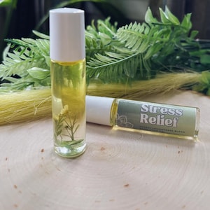 May include: Two clear glass roll-on bottles with white caps. One bottle contains a yellow liquid with small green plants inside. The other bottle has a label that reads "Stress Relief" in white text on a green background. The bottles are on a wooden surface.