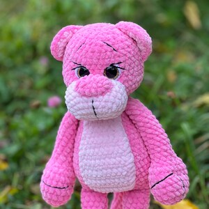 May include: A pink crocheted plush toy shaped like a panther with large brown eyes. The toy has a pink and white striped body and black embroidered details.