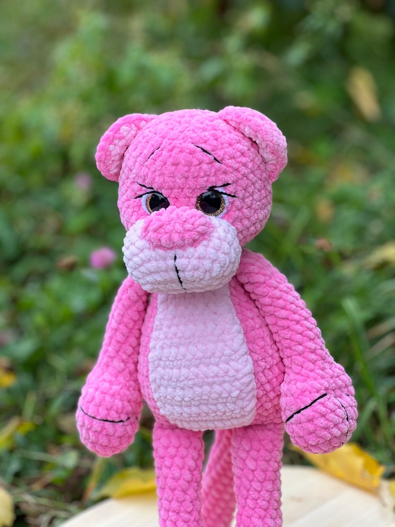 May include: A pink crocheted plush toy shaped like a panther with large brown eyes. The toy has a pink and white striped body and black embroidered details.