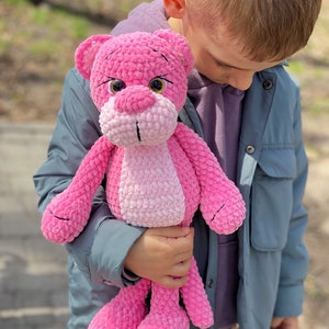 May include: A child holds a pink crocheted stuffed animal that resembles a panther. The stuffed animal has black eyes and a pink nose.