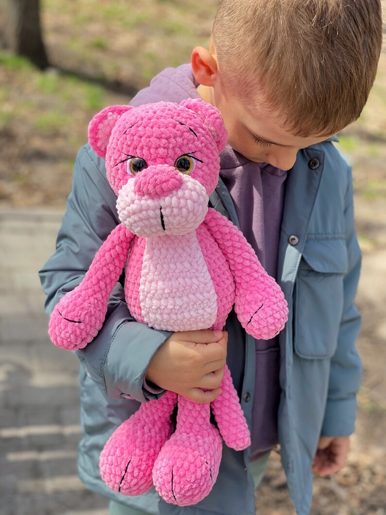 May include: A child holds a pink crocheted stuffed animal that resembles a panther. The stuffed animal has black eyes and a pink nose.