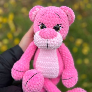 May include: Pink crocheted plush toy shaped like a panther with black eyes and a white nose. The toy is being held by a person.