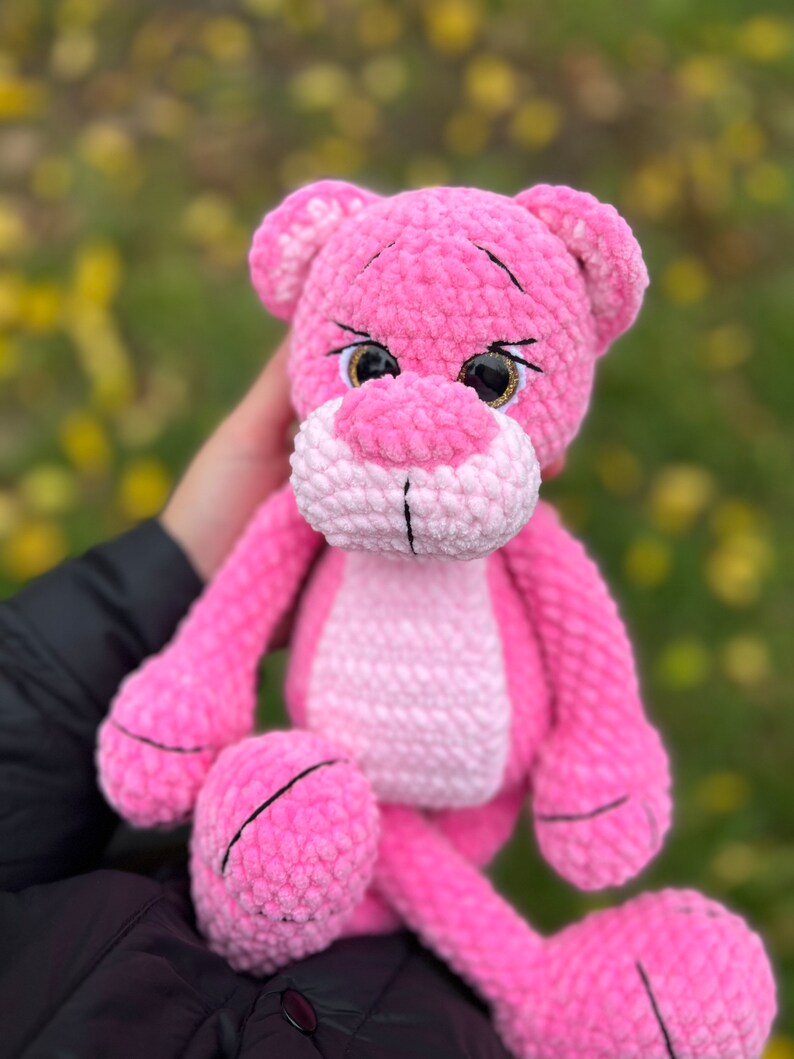 May include: Pink crocheted plush toy shaped like a panther with black eyes and a white nose. The toy is being held by a person.