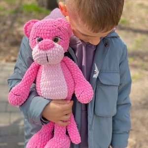 May include: A child holds a pink crocheted stuffed animal that resembles a panther. The stuffed animal has a pink and white ombre effect and black embroidered eyes.