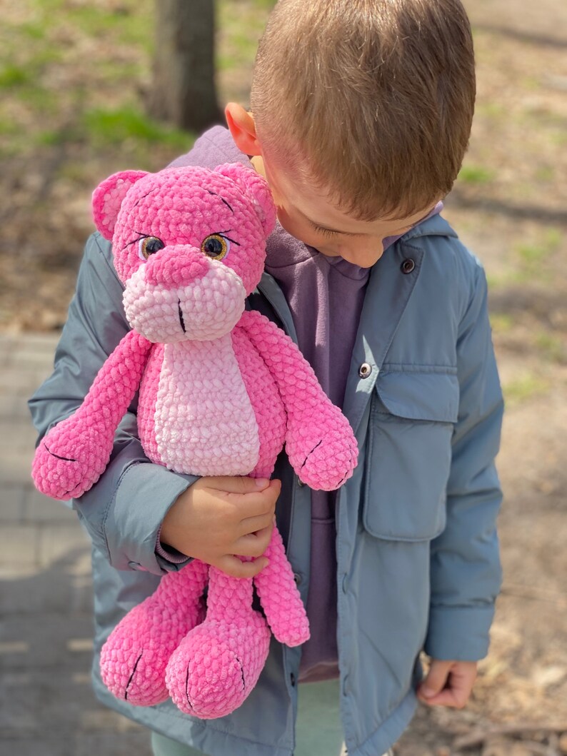 May include: A child holds a pink crocheted stuffed animal that resembles a panther. The stuffed animal has a pink and white ombre effect and black embroidered eyes.
