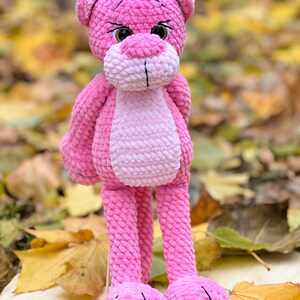 May include: A pink crocheted plush toy shaped like a panther with long legs and large eyes. The toy is sitting on a wooden surface with fall leaves around it.