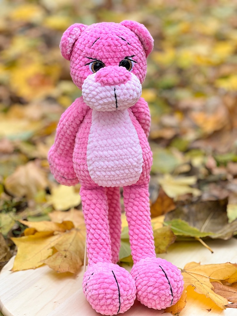 May include: A pink crocheted plush toy shaped like a panther with long legs and large eyes. The toy is sitting on a wooden surface with fall leaves around it.