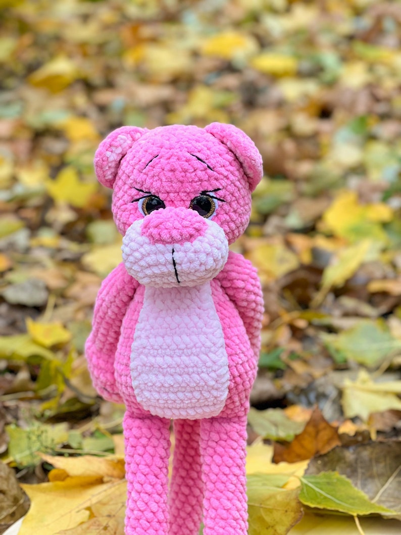 May include: A pink crocheted plush toy shaped like a bear with large brown eyes. The bear is standing on a bed of fallen autumn leaves.