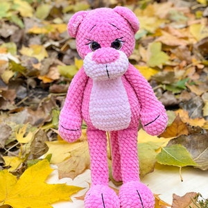 May include: A pink crocheted plush toy shaped like a bear with large brown eyes. The bear is standing on a wooden surface with autumn leaves scattered around it.