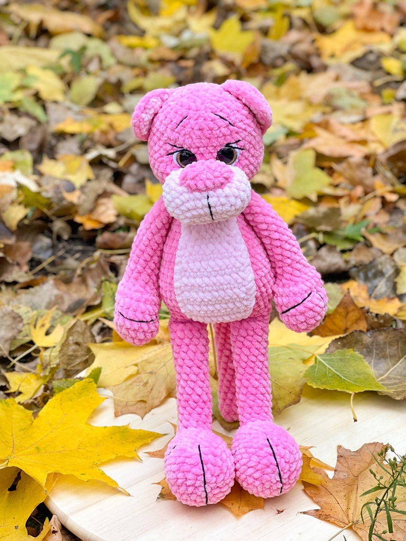 May include: A pink crocheted plush toy shaped like a bear with large brown eyes. The bear is standing on a wooden surface with autumn leaves scattered around it.