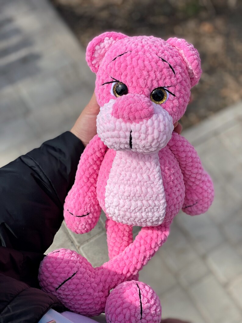 May include: Pink crocheted plush toy shaped like a bear with large, sparkly eyes. The bear has long, thin legs and a pink and white ombre color scheme.