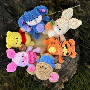 May include: A collection of crocheted stuffed toys, including a blue donkey, a tan bunny, a yellow and red Winnie the Pooh, a pink pig, a brown bear, and an orange tiger. The toys are arranged in a circle on a black wire rack.