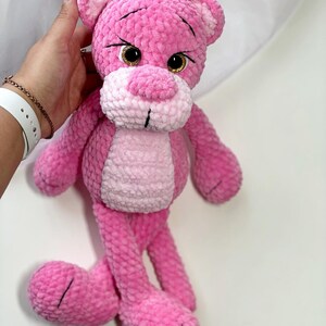 May include: A pink crocheted plush toy in the shape of a bear with large brown eyes. The bear has a pink and white striped body and long pink legs.