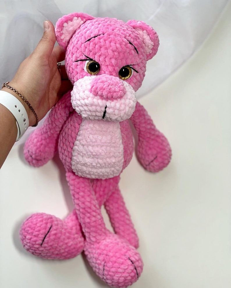 May include: A pink crocheted plush toy in the shape of a bear with large brown eyes. The bear has a pink and white striped body and long pink legs.