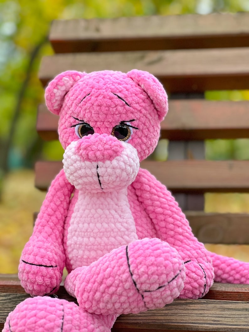 May include: A pink crocheted plush toy in the shape of a panther sitting on a wooden bench. The toy has black eyes and a pink and white ombre effect.