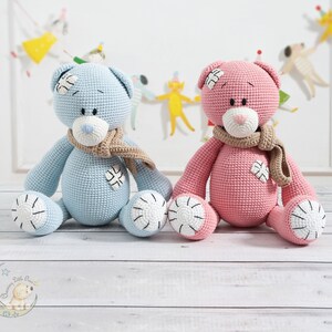 May include: Two crocheted teddy bears, one blue and one pink, are sitting on a white wooden surface. Both bears have a brown scarf and a white patch on their chest.