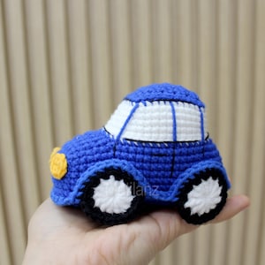 Handmade crocheted toy car in vibrant blue with white roof and wheels, featuring a yellow front light, crafted from soft yarn for a cozy texture.
