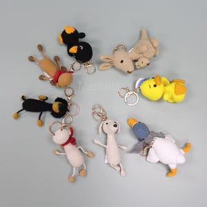 May include: A collection of handmade crochet animal keychains in various colors. The keychains include a black and yellow penguin, a brown dog, a yellow duck, a white dog, a white goose, and a black dog. Each keychain has a gold-colored ring.