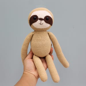 Könnte beinhalten: Handgefertigte Häkeltierpuppe in Beige- und Cremetönen. Das Faultier hat einen runden Kopf mit braunen Augenpartien und schwarzen Knopfaugen. Die langen Arme und Beine sind schlank. Die Puppe wird in einer Hand gehalten.