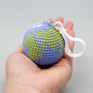 May include: A crocheted Earth keychain, featuring blue oceans and green continents. The globe is held in a hand, with a white clip attached. The keychain is a small, round, and colorful representation of the planet.