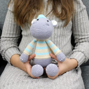 Handmade crocheted hippo toy in soft gray with light blue and cream striped details, featuring a cozy knit hat and textured ears.