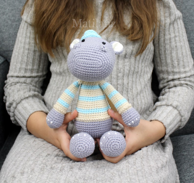 Handmade crocheted hippo toy in soft gray with light blue and cream striped details, featuring a cozy knit hat and textured ears.