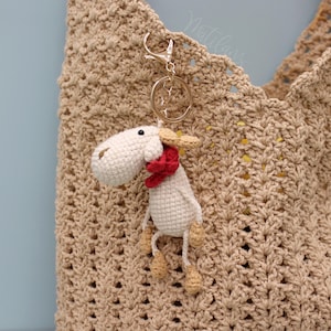 May include: A handmade crochet sheep keychain with a red scarf and tan hooves hangs from a gold-colored clasp. The sheep is attached to a tan crochet bag with a decorative open weave pattern. The background is a light blue.