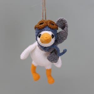 May include: A crocheted white goose ornament with orange feet and a yellow beak. The goose wears a blue aviator cap with brown goggles and a grey scarf. A brown twine string is attached to the top of the cap for hanging the small stuffed figure.