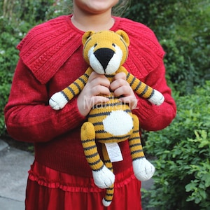 May include: A child holds a knitted tiger plush toy. The toy is yellow and black striped with white accents. The child is wearing a red sweater with a large collar.