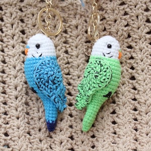 May include: Two crochet parakeet keychains. One is blue with a white head and orange beak, the other is green with a white head and orange beak. Both have black eyes and are attached to gold key rings. The background is a beige crocheted fabric.