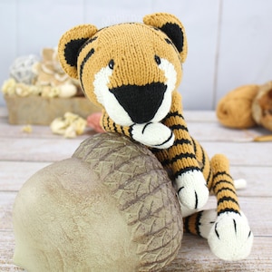 May include: A knitted tiger plush toy with a tan body, black stripes, and white accents. The tiger has a black and white face and is posed with its paws resting on a decorative acorn-shaped object. The background is a light wood surface.