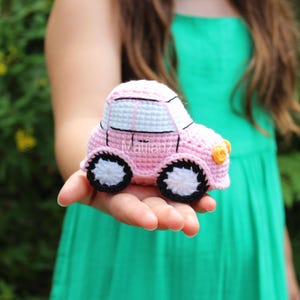 Handmade crocheted car toy in soft pink with white accents, black wheels, and a yellow front bumper, crafted from yarn for playful charm.