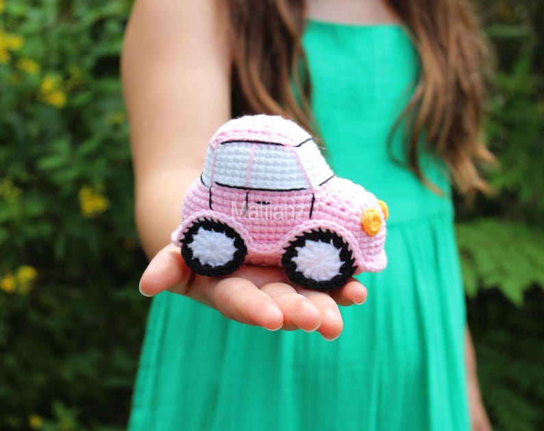 Handmade crocheted car toy in soft pink with white accents, black wheels, and a yellow front bumper, crafted from yarn for playful charm.