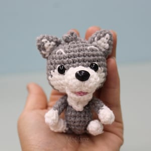 May include: A handmade crochet wolf figurine in gray and white, with black eyes and a pink mouth. The wolf is held in a hand, showcasing its small size and detailed craftsmanship. The figurine is a cute, soft toy.