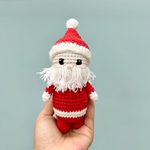 May include: A handmade crochet Santa Claus doll. The doll features a red suit with white trim, a white beard, and a red hat with a white pom-pom. The eyes are black, and the doll is held in a hand against a light blue background.